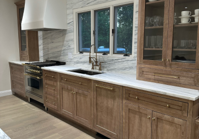 Medium-brown inset cabinets with furniture-style tall glass wall cabinets sitting on a white stone countertop with an artisan white kitchen hood