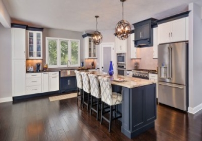 Open white shaker kitchen with black crown molding and toe kick with large black island offering seating for four, ball pendant island lighting, wood range hood over gas cooktop, and glass door display cabinets next to sink window.