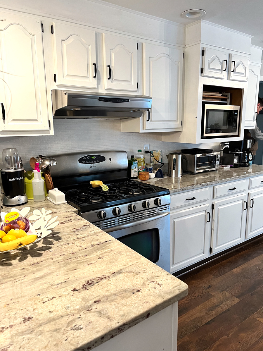 Client's before kitchen photo with partial overlay cabinets with white arched raised panel doors, exposed black hinges, soffits over the wall cabinets, and an awkward layout Client's before kitchen photo with partial overlay cabinets with white arched raised panel doors, exposed black hinges, soffits over the wall cabinets, and an awkward layout