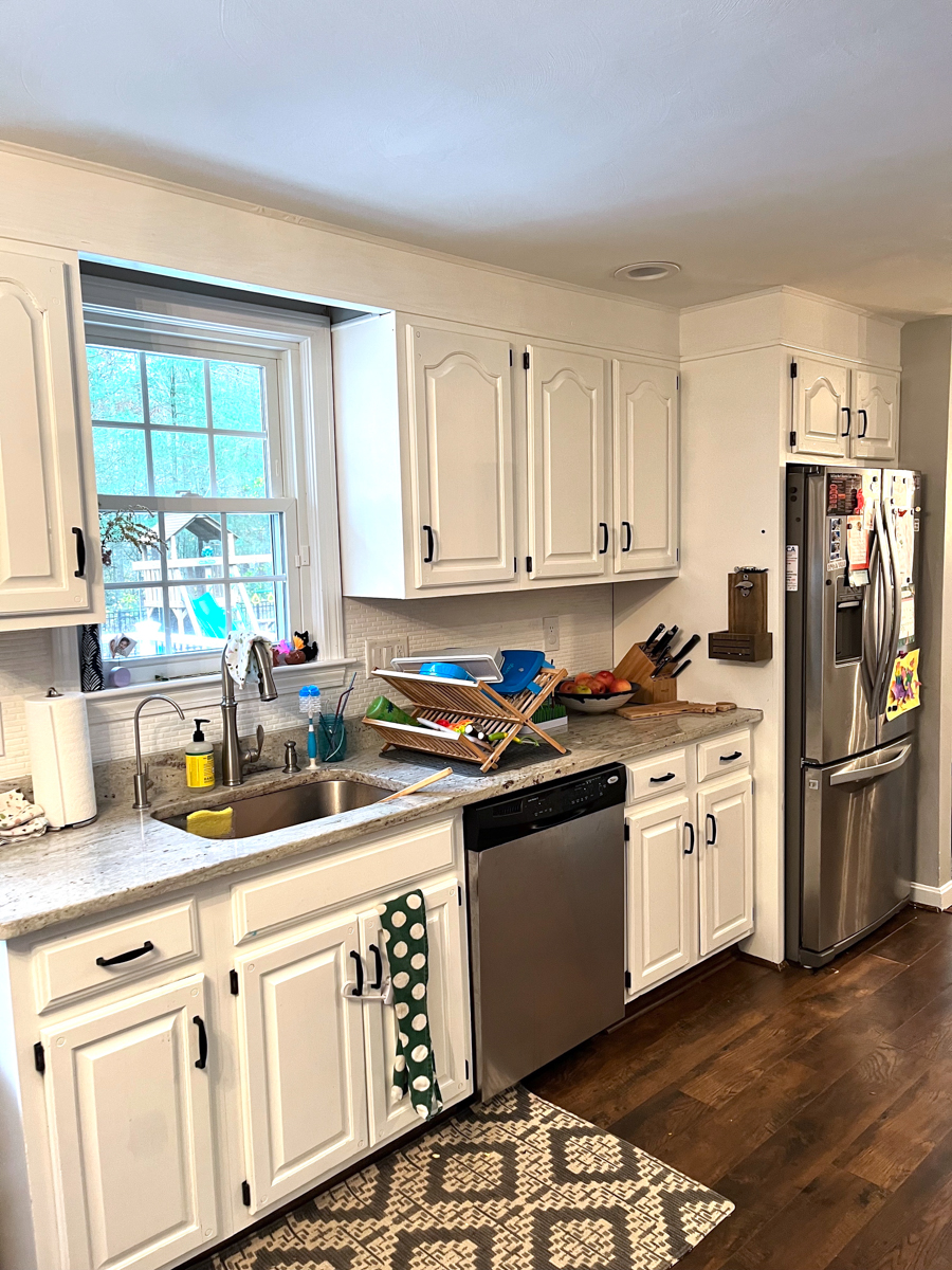 Client's before kitchen photo with partial overlay cabinets with white arched raised panel doors, exposed black hinges, soffits over the wall cabinets, and an awkward layout Client's before kitchen photo with partial overlay cabinets with white arched raised panel doors, exposed black hinges, soffits over the wall cabinets, and an awkward layout