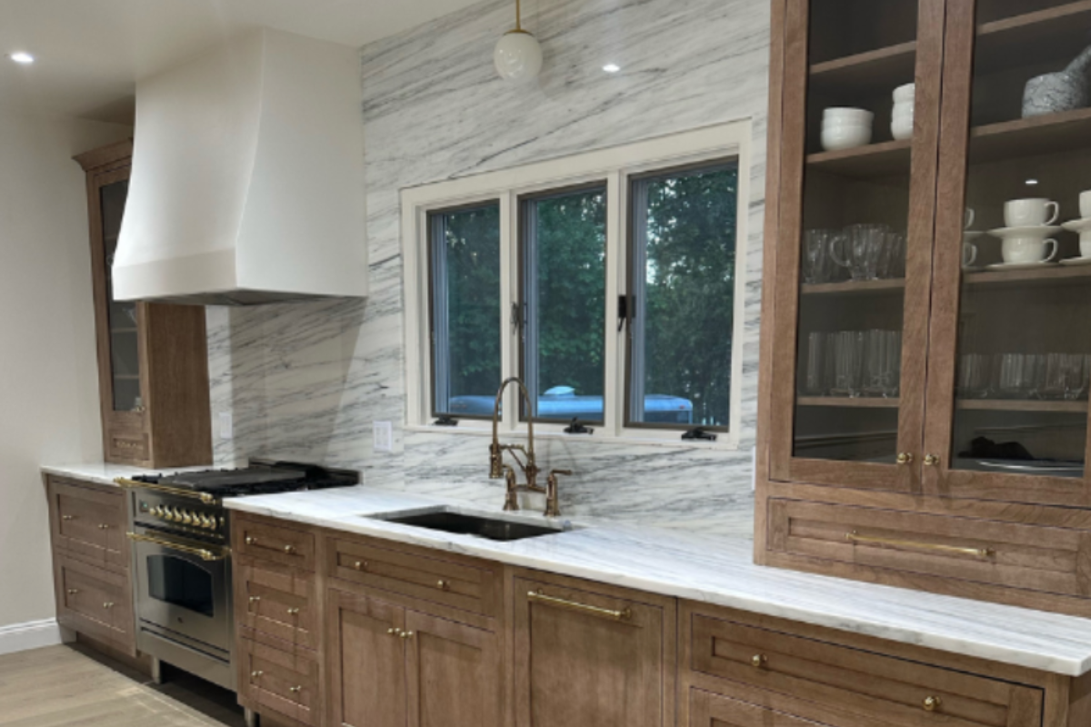 Medium wood tone inset kitchen cabinets with a gas range, white plaster hood and white stone slab covered wall Medium wood tone inset kitchen cabinets with a gas range, white plaster hood and white stone slab covered wall