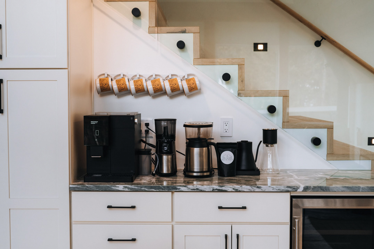 White shaker coffee bar cabinets next to a glass staircase railing with black cabinet pulls and coffee cups above White shaker coffee bar cabinets next to a glass staircase railing with black cabinet pulls and coffee cups above