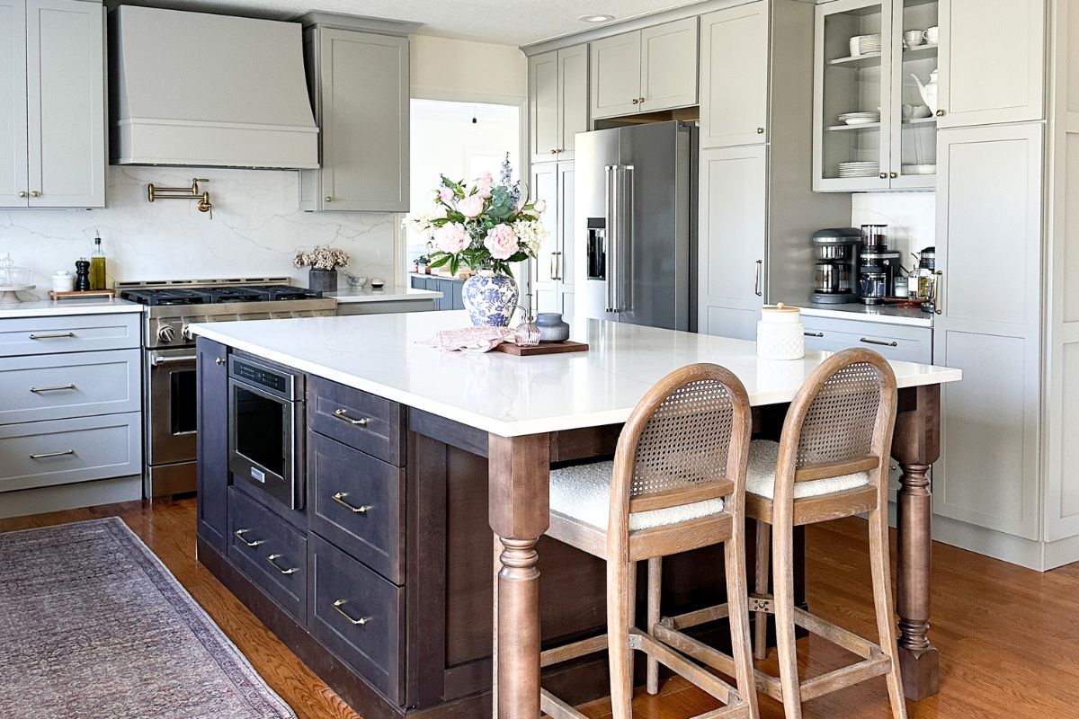 Light gray shaker kitchen with wood hood, white quartz countertops, and a large dark brown furniture-style island Light gray shaker kitchen with wood hood, white quartz countertops, and a large dark brown furniture-style island