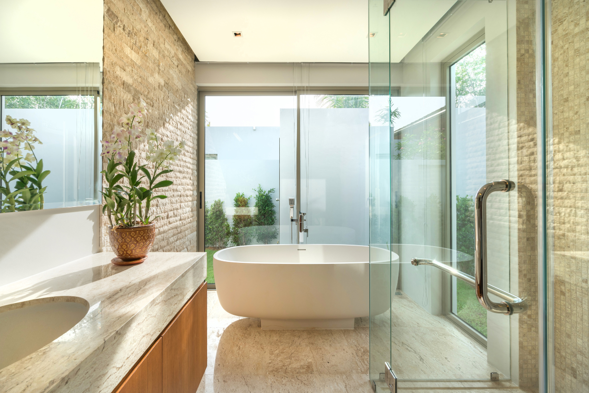 Large organic bathroom with wood vanity cabinet and concrete countertop with freestanding tub in front of a large window next to focal wall in natural stone Large organic bathroom with wood vanity cabinet and concrete countertop with freestanding tub in front of a large window next to focal wall in natural stone