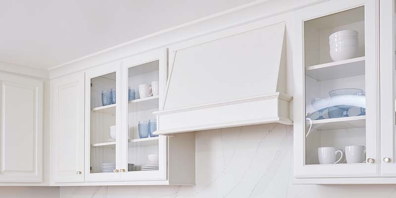 CliqStudios Decorative cabinet in Painted White with glass doors displaying glassware and dishware and a wood range hood in the Classic style CliqStudios Decorative cabinet in Painted White with glass doors displaying glassware and dishware and a wood range hood in the Classic style