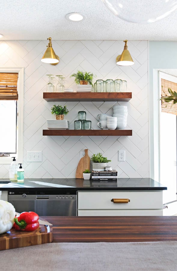 Floating shelves with glassware and plants against white subway tile backsplash in a herringbone pattern. Floating shelves with glassware and plants against white subway tile backsplash in a herringbone pattern.