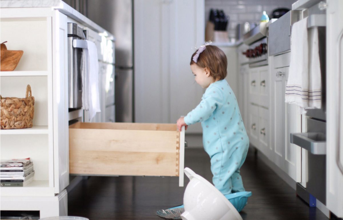 A baby in a blue onsie opening a lower cabinet drawer. A baby in a blue onsie opening a lower cabinet drawer.