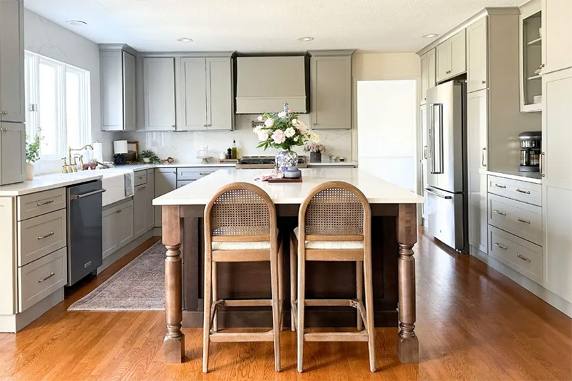 Duluth Pebble & Newport Chai kitchen cabinets Large Farmhouse-style kitchen with light gray shaker cabinets and a dark wood center island and wicker counter stools and white marble-like quartz countertops.