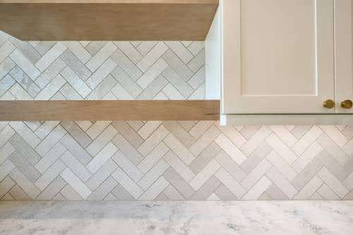 White shaker cabinets with a light cream herringbone tile backsplash and light wood floating shelves White shaker cabinets with a light cream herringbone tile backsplash and light wood floating shelves